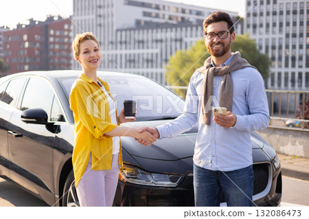A man and a woman shake hands in front of a black electric car, possibly after a business deal. 132086473