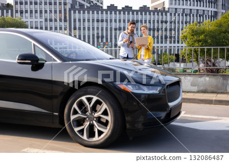 A man and woman discuss work while standing near a sleek black electric car in an urban setting. 132086487