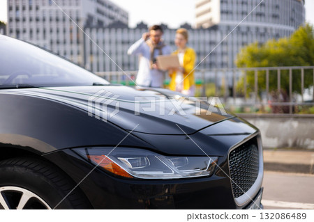 A sleek black car is the focus, with a blurred couple in the background near a city building. A sleek black car is the focus, with a blurred couple in the background near a city building. 132086489