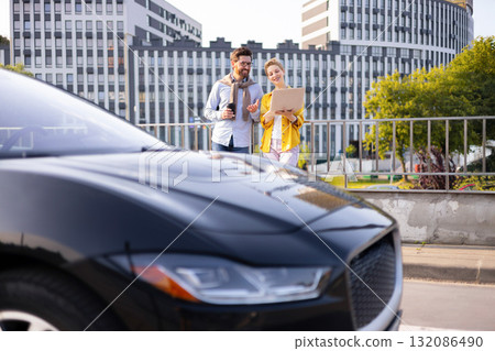 Two business colleagues are discussing a project while standing near a modern black car in an urban setting. 132086490