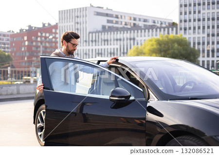A man in glasses opens the door of a sleek, black car, likely preparing to drive in an urban setting. 132086521