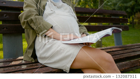 Pregnant holding a book in hands while on morning walk having a rest. Pregnant holding a book in hands while on morning walk having a rest. 132087311