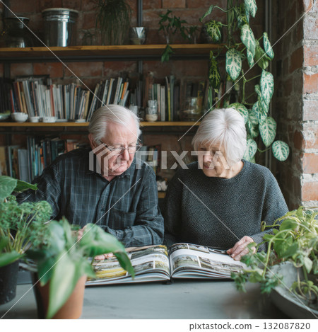 An elderly couple sits closely, looking through a photo album or magazine in their cozy, plant-filled home with exposed brick and a bookshelf. The scene portrays retirement, leisure, shared memories 132087820