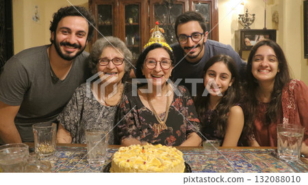 A close-up, authentic portrait of a multi-generational family celebrating a birthday indoors. They are all smiling happily behind a yellow cake, with the grandmother wearing a party hat. 132088010
