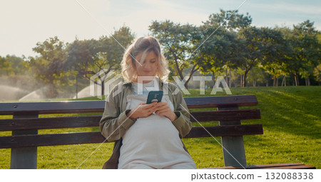 Smiling pregnant woman using phone and chatting outside. Smiling pregnant woman using phone and chatting outside. 132088338