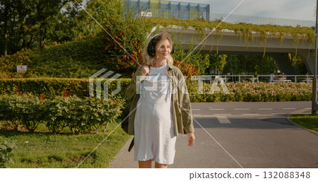 Pregnant woman with headphones on her head walks in the park and listens to music. 132088348