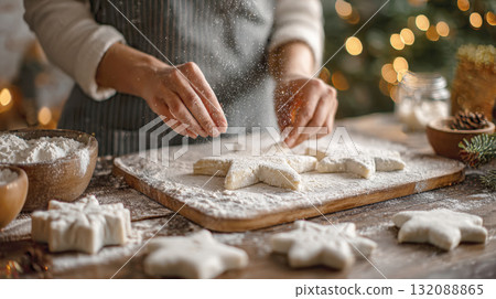 A close-up of a person's hands sprinkling flour over star-shaped cookie dough cutouts on a board, with a christmas tree and lights in the background. Captures holiday baking, cozy winter atmosphere 132088865