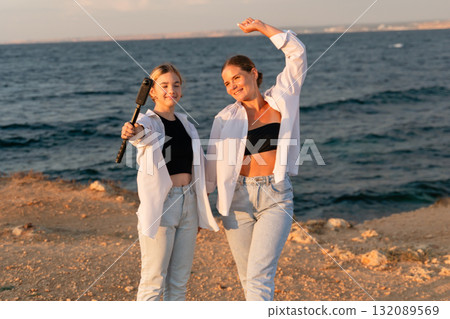 Women Beach Sunset Selfie - Two women take a selfie on a beach at sunset, one holding a selfie stick. Women Beach Sunset Selfie - Two women take a selfie on a beach at sunset, one holding a selfie stick. 132089569
