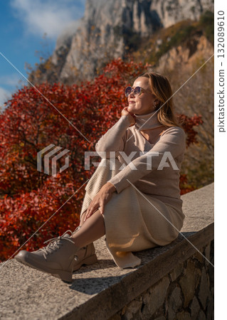 Woman Autumn Mountainside Contemplation: A woman sits contemplatively on a stone wall overlooking a vibrant autumnal mountain landscape, enjoying the scenic view. Woman Autumn Mountainside Contemplation: A woman sits contemplatively on a stone wall overlooking a vibrant autumnal mountain landscape, enjoying the scenic view. 132089610