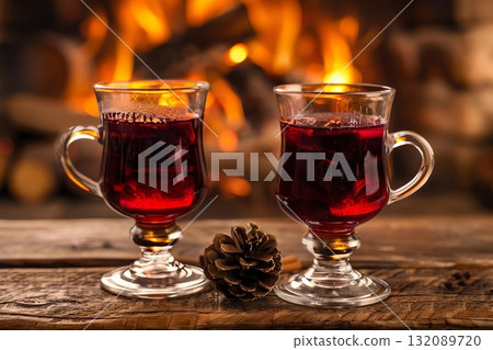 Two elegant glass mugs filled with steaming red beverage sit on a rustic wooden table, with a cozy fireplace glowing warmly in the background, creating a festive atmosphere 132089720
