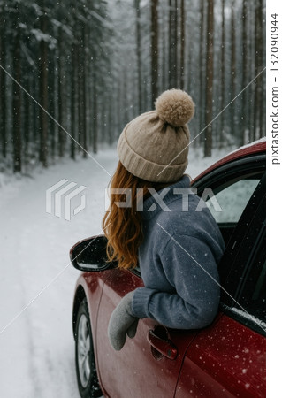 Winter road trip through snowy forest with woman in cozy hat leaning from red car 132090944