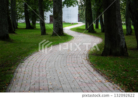 Curved brick walkway winding through green park with tall trees and leading towards a white building Curved brick walkway winding through green park with tall trees and leading towards a white building 132092622