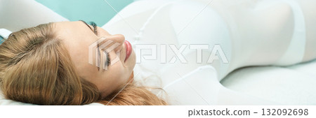 Relaxed young caucasian female in white outfit lying on bed with soft 132092698