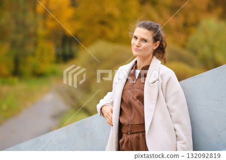 Young woman poses casually outdoors in autumn landscape with colorful leaves and gray structure 132092819