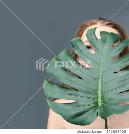 Caucasian female young adult partially concealed by large monstera leaf against Caucasian female young adult partially concealed by large monstera leaf against 132092908