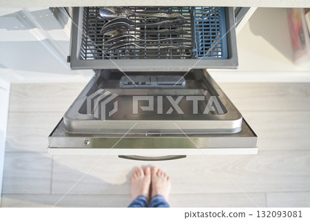 Open dishwasher with utensils tray and person standing barefoot on light wooden 132093081