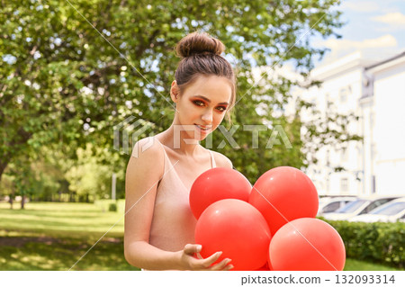 Young caucasian female holding red balloons in sunny park setting with greenery 132093314