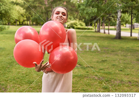 Smiling young caucasian female holding red balloons in a park on a sunny day Smiling young caucasian female holding red balloons in a park on a sunny day 132093317