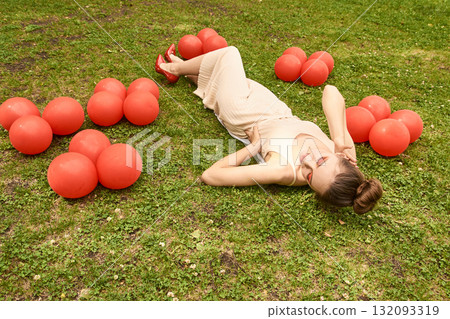 Young caucasian female in beige dress lying on grass surrounded by red balloons 132093319