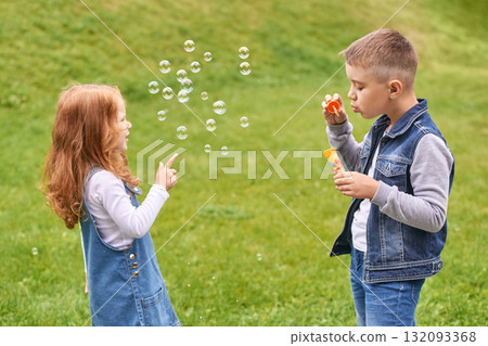 Young caucasian children blowing bubbles outdoors on grass in denim outfits 132093368