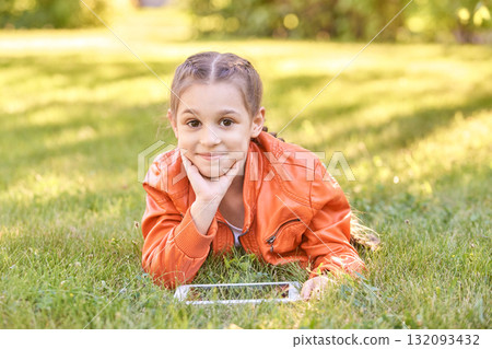 Smiling caucasian female child lying on grass in orange jacket with tablet Smiling caucasian female child lying on grass in orange jacket with tablet 132093432