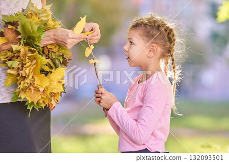 Caucasian young girl holding autumn leaves talking to adult female outdoors 132093501