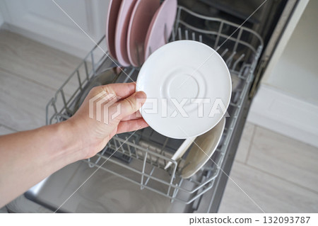 Caucasian female young adult loading dishwasher with clean white saucer and pink Caucasian female young adult loading dishwasher with clean white saucer and pink 132093787