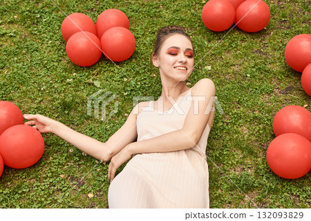 Young caucasian woman relaxing on grass with red balloons, enjoying outdoor Young caucasian woman relaxing on grass with red balloons, enjoying outdoor 132093829