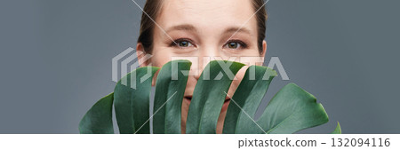 Caucasian adult woman smiling behind monstera leaf against gray background Caucasian adult woman smiling behind monstera leaf against gray background 132094116