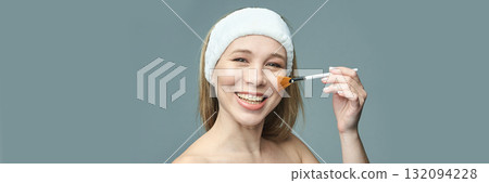 Smiling caucasian female using makeup brush in skincare routine on blue Smiling caucasian female using makeup brush in skincare routine on blue 132094228
