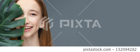 Smiling young caucasian female behind monstera leaf on grey background Smiling young caucasian female behind monstera leaf on grey background 132094282