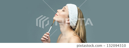 Young caucasian female with headband applying makeup with brush on chin, side Young caucasian female with headband applying makeup with brush on chin, side 132094310