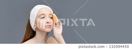 Young caucasian female cleansing face with sponge and headband on gray 132094450