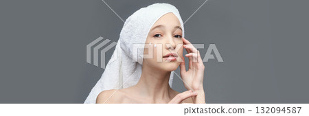 Young caucasian female applying skincare cream with towel on head against gray 132094587