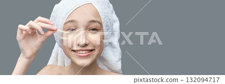 Young caucasian female applying eye patches smiling in towel headwrap on gray Young caucasian female applying eye patches smiling in towel headwrap on gray 132094717