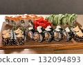 A delicious, high-angle close-up shot of a large, colourful assortment of sushi rolls presented on a rustic wooden board, showcasing a variety of textures and ingredients 132094893