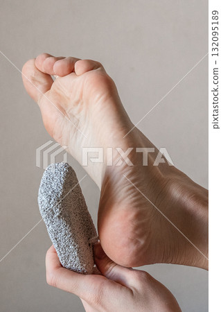 A close-up of a hand using a pumice stone on a foot. The foot is bare, showing smooth skin and a well-groomed heel. The background is neutral and simple. 132095189