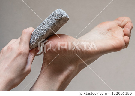 A close-up of a hand using a pumice stone on a foot. The foot is bare, showing smooth skin and a well-groomed heel. The background is neutral and simple. A close-up of a hand using a pumice stone on a foot. The foot is bare, showing smooth skin and a well-groomed heel. The background is neutral and simple. 132095190