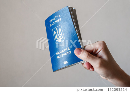 A hand holding a blue Ukrainian passport. The passport features the national emblem and text in Ukrainian and English. The background is neutral and simple. A hand holding a blue Ukrainian passport. The passport features the national emblem and text in Ukrainian and English. The background is neutral and simple. 132095214
