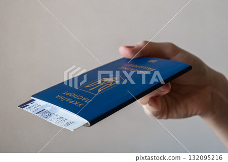 A hand holding a blue Ukrainian passport. The passport features the national emblem and text in Ukrainian and English. The background is neutral and simple. A hand holding a blue Ukrainian passport. The passport features the national emblem and text in Ukrainian and English. The background is neutral and simple. 132095216