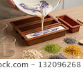A close-up shot of a person pouring natural oils from small glass beakers into a creamy white soap base in a wooden bowl, then stirring the mixture with a wooden spoon to blend the ingredients 132096268