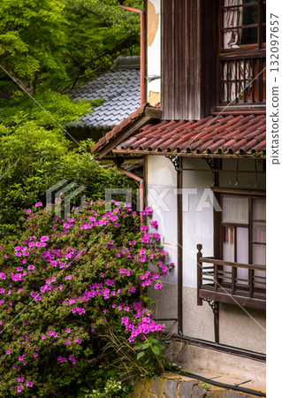 Traditional Japanese countryside house with a tiled roof surrounded by greenery and blooming pink flowers Traditional Japanese countryside house with a tiled roof surrounded by greenery and blooming pink flowers 132097657