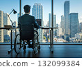 A young man sits in a wheelchair in front of a computer desk in an office 132099384