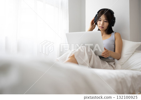 一位年輕女子戴著耳機在床上操作筆記型電腦 132100925