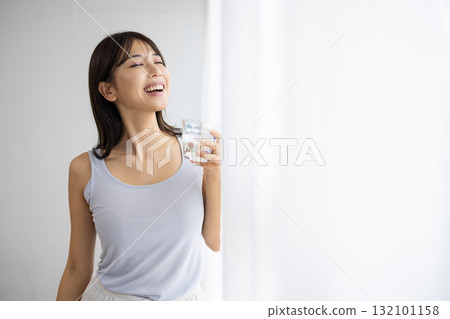 Smiling young woman holding a glass of water by the window Smiling young woman holding a glass of water by the window 132101158