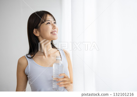 Smiling young woman holding a bottle of water by the window Smiling young woman holding a bottle of water by the window 132101216