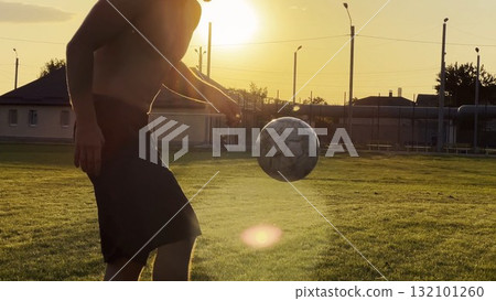 Young man juggling soccer ball on stadium at sunset. Professional footballer kicking ball at green field. Sportsman practicing tricks at meadow with sunlight at background. Freestyle football Young man juggling soccer ball on stadium at sunset. Professional footballer kicking ball at green field. Sportsman practicing tricks at meadow with sunlight at background. Freestyle football 132101260
