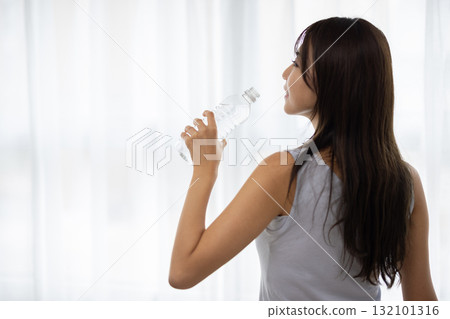Rear view of a young woman drinking bottled water by the window 132101316