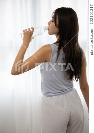 一位年輕女子在窗邊喝瓶裝水的後視圖 132101317
