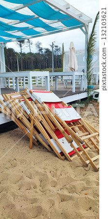 Neatly stacked red and white wooden lounge chairs on a sandy terrace next to a beach hut. Concept of the end of the beach season, a small business in operation 132103621